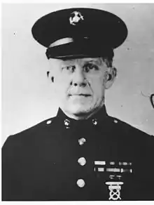 Head and shoulders of a white man wearing a peaked cap and a dark military jacket with ribbon bars on the left breast and bright buttons down the center.