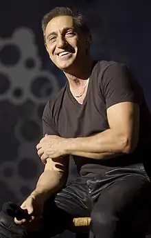 An image of a man wearing a black shirt and pants is sitting on a stool with a microphone on his right hand