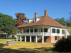 A two story house with a large porch upstairs, two chimneys, and a lot of trees behind it.