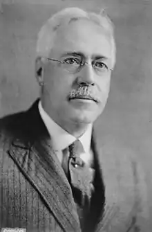 An old man with grey hair and a moustache, wearing a suit and glasses