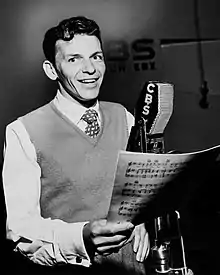 Photograph of a man wearing a shirt, tie, and sweater vest, holding sheet music, singing into a microphone