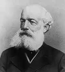 A black and white image of a bald man in a dark outfit, with a bushy white beard and mustache