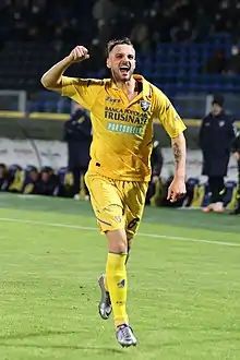 A smiling man wearing a yellow sports shirt running on a football field holding a bent arm