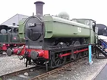 A pannier tank locomotive is seen from the front a slightly to the right. The cab and pannier tank are painted green. The chimney, smokebox, running plate, splashers, and wheels are black, and the buffer beam and coupling rods are red. The number, 7754, is shown in white letters on a black plate on the smokebox door. Part of round cab window is visible and is edged in brass. The white lettering "NCB 7754" is on the side of the pannier tank.