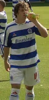 A footballer wearing a white and blue hooped football shirt and shorts. He is standing on the pitch and drinking from a bottle.