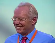 A grinning older white man with receding white hair and glasses, wearing a blue dress shirt with red necktie and an orange strap around his neck, seen from just below the neck up, looks to the camera's left on a green background