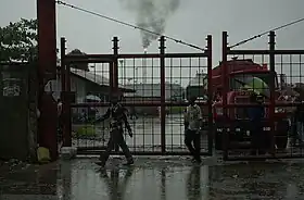 A picture of gates of the Port Harcourt Refining Company Ltd. with an oil refinery's fire seen in the background in rainy weather.