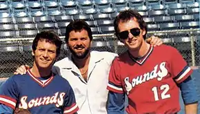 Three men standing in front of blue stadium seats. One is wearing a blue baseball jersey, one is in a white dress shirt, and one is in a red baseball jersey.