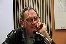 Head and shoulders of a man wearing a green t-shirt and black leather jacket. His hair is thinly cropped looks like he's contemplating something.