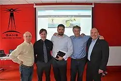 Five smiling men standing in front of a wall with the GANS logo