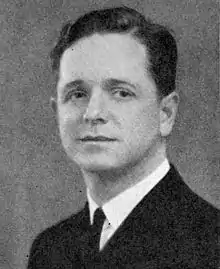 Head and shoulders of a white man with carefully combed and parted wavy hair, wearing a dark jacket over a shirt and tie.