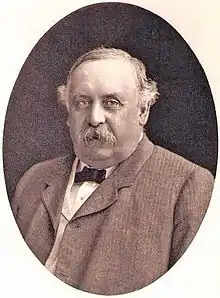 A sepia-toned oval portrait of a mustached man with a jacket and bowtie