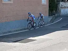 Two cyclists, both wearing white and blue jerseys, one with yellow trim, ride on an empty road beside a red and blue building.