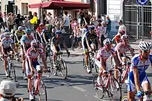 A group of cyclists riding on the road. Spectators watch at the roadside.