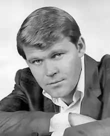 A black and white portrait of a man smiling with his arms crossed, staring into the lens of the camera