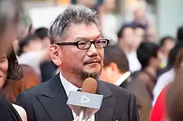 A bespectacled Hideaki Anno in front of a microphone