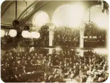 A photograph showing a large number of men seated on semi-circular tiers in a vaulted chamber as a large crowd looks on from an arcaded balcony