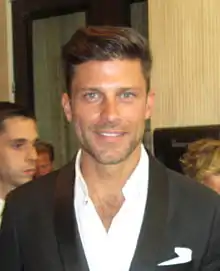 A man with dark hair wearing a white T-shirt including a silver necklace.