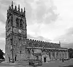 A church seen from the southwest with the tower on the left and the body of the church, with clerestory and south aisle, to the right. The tower has crocketted pinnacles and the body of the church is crenellated