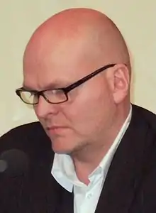 bald white male in profile, wearing glasses, white shirt and dark jacket, looking down, with a microphone in front