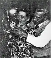 Black and white photograph of a man standing behind a woman in a flowered dress, both of whom have their hands raised to eye-level while holding a camera