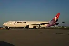 A white twin-engine plane painted with the word "HAWAIIAN" in the front above the windows, a black, silver, and white logo of a pirate's head on the forward bottom of the fuselage, and a woman in different purple hues on the tail taxis at an airport