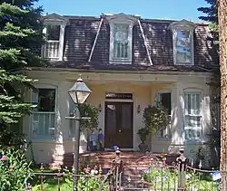 A two-story house of painted beige brick on the first story with a steeply-angled dark brown shingled roof on top from which three dormer windows project, the middle one from a slightly projecting trapezoid. The ground floor has two identical projecting bay windows, with a double wooden door in the middle atop a short flight of brick steps. Flowerpots hang from the roof and there are two small figurines on the porch. In front is an iron fence, lamppost, and flowers. At the right is part of a tree trunk, with part of an evergreen on the left.