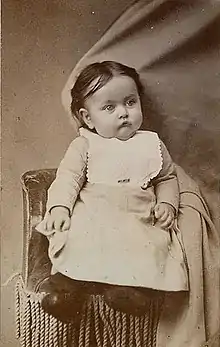 A toddler in a dress and bib, head cradled by a curtain disguising an arm