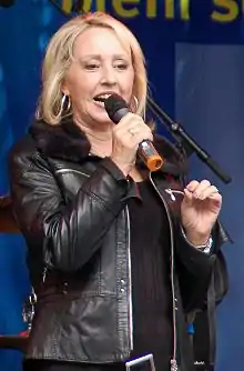 Hildegard Krekel standing onstage wearing black, speaking into a microphone