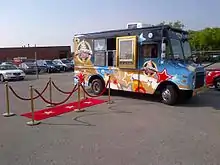  Blue-and-gold van with a window for selling food out of the side with chalkboard menus and a red carpet leading up to the window