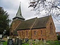 A red sandstone church seen from the southeast. The chancel is slightly lower than the nave and the tower has a broach spire. Gravestones are in the foreground and part of a tree is to the left