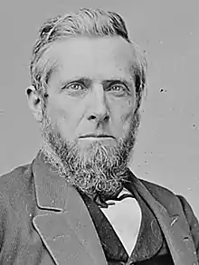 A man with white hair and a white beard and mustache wearing a white shirt, black jacket and black vest