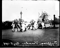 Black-and-white photo of horses pulling a parade float down a city street.