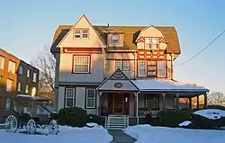 An intricate three-story beige wooden house with red trim, with snow on the porch roof and the front lawn, with a setting sun lighting the upper stories from the right. It has windows of different shapes and sizes. A front porch is on the right half of the first story, with a triangular projection on the left with a prominent "36" in the center. Another triangular section projects from the left hand side of the roof, with a smaller projection with a segmented, more curvilinear roofline to its right. A brick building is visible on the left; in the left front of the yard is an old horse-drawn carriage.