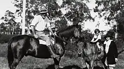 A man on a horse at left looking at camera with a small boy on another horse and a woman standing next to the boy