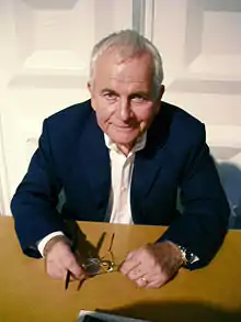 A color photograph of a man in a suit, sitting at a brown desk, holding a pair of glasses