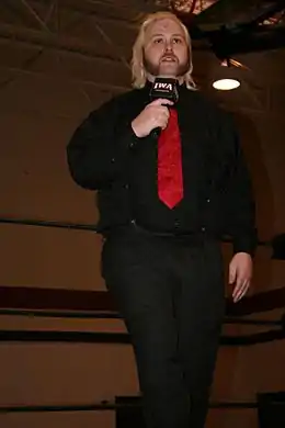 A male wrestler dressed casually in a wrestling ring holding a microphone