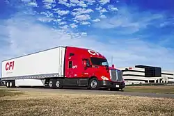 Tractor-trailer in front of large, multi-story office building, CFI headquarters in Joplin. Tractor is painted red with a white CFI logo on the face of the roof fairing. Trailer is painted white with a red CFI logo on the side near the tail.