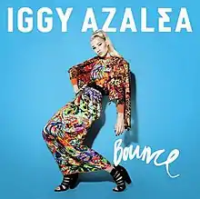 A portrait coloured in light blue, featuring a young blonde woman at its forefront. She is posing in a crouched standing position with her one hand on her hip. She is wearing a traditional Indian sari composed of various colours, and her hair is styled in a high ponytail. Above her in bold white capital font is the name 'Iggy Azalea'. Below her loose arm, in thin cursive white font is the title 'Bounce'.