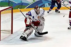 Ilya Bryzgalov is on the ice deflecting a hockey puck. He is wearing a white and blue jersey with red pants.