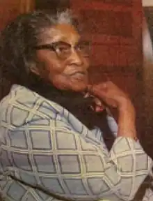 Black woman in a checked jacket wearing glasses with her right thumb on the left part of her chin