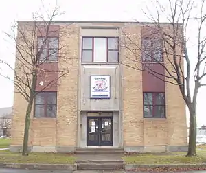  A two-story brick building with four large windows evenly spaced, and a large logo in between them, like the number five on a die, two leafless trees flank the building's entrance.