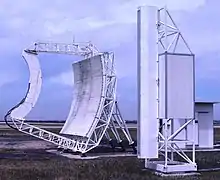 Colour photograph of two large white-painted items of equipment (the left being a parabolic 'dish' with straight sides; the right a tall, narrow structure) on grass, with cloudy sky above.