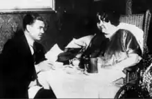 A man and a woman, both white, seated at a table set for dining; the man is wearing a suit; the woman is wearing a dress and sitting in a wicker wheelchair