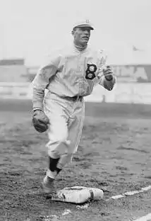 A man in a light baseball uniform with a dark "B" on the left chest running past first base with a fielding glove on his right hand