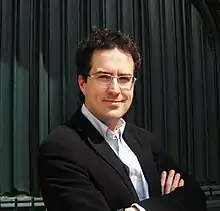 James Hannigan standing against a curtain backdrop.  He is wearing a suit and glasses, and stands with his arms folded and a smile.