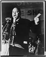 James Farmer, three-quarter length portrait, speaking before microphones outside Hotel Theresa