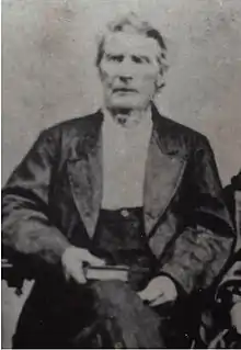 An elderly man is seated with a book in his right hand in his lap