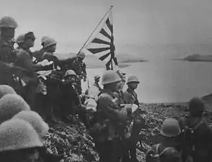 alt=Historic photograph of Japanese troops raising the Imperial battle flag on Kiska Island.