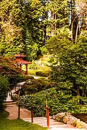 There are tall trees in the background. There is a gazebo below it. There is a walkway leading up to the gazebo. There are trees on both sides of the walkway.